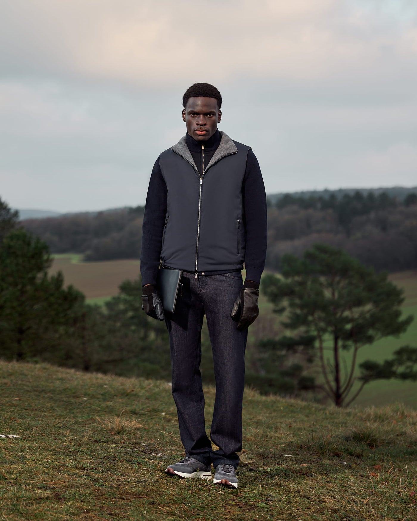 Man standing outdoors in a natural landscape wearing a dark jacket and pants.