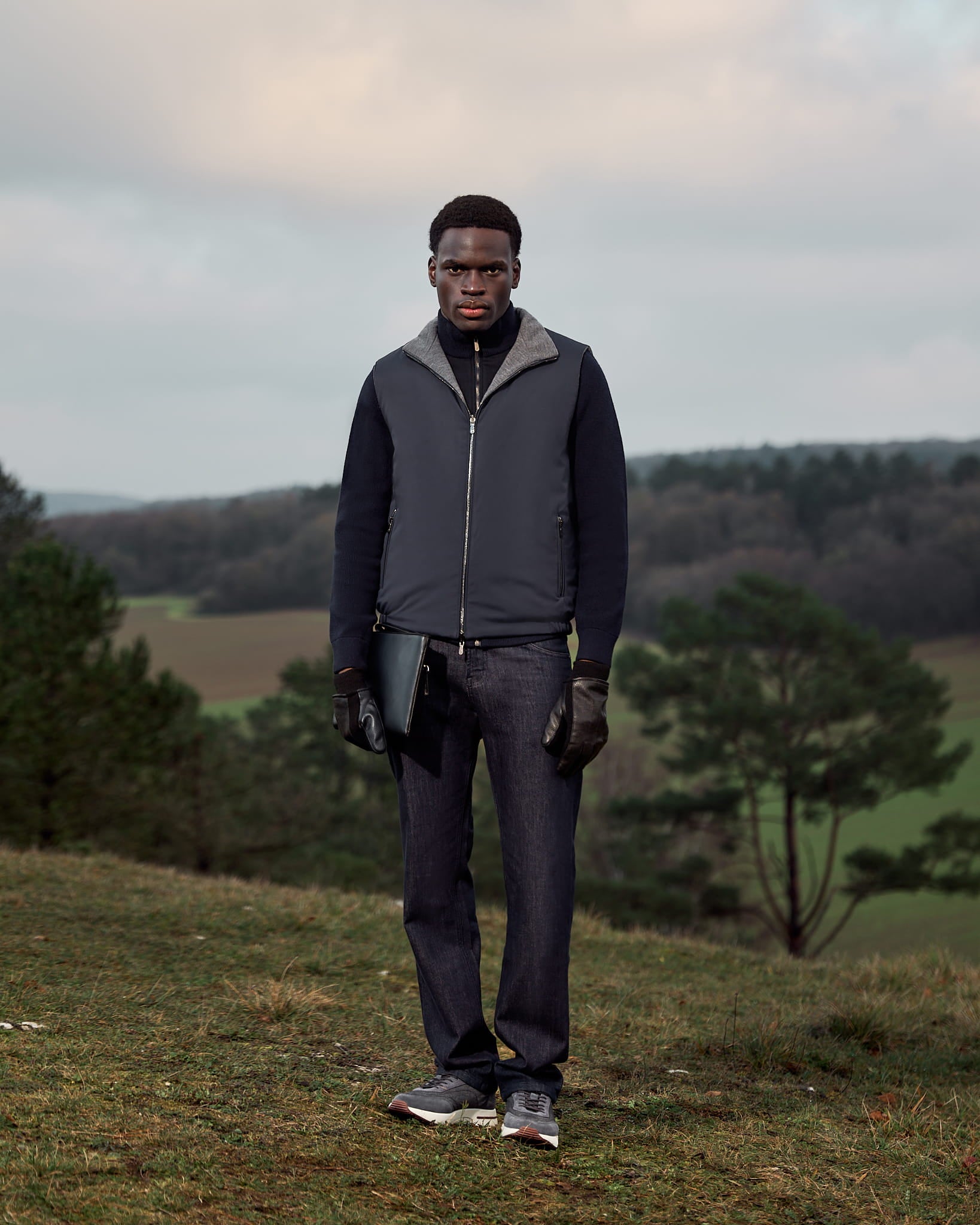Man standing outdoors in a natural landscape wearing a dark jacket and pants.