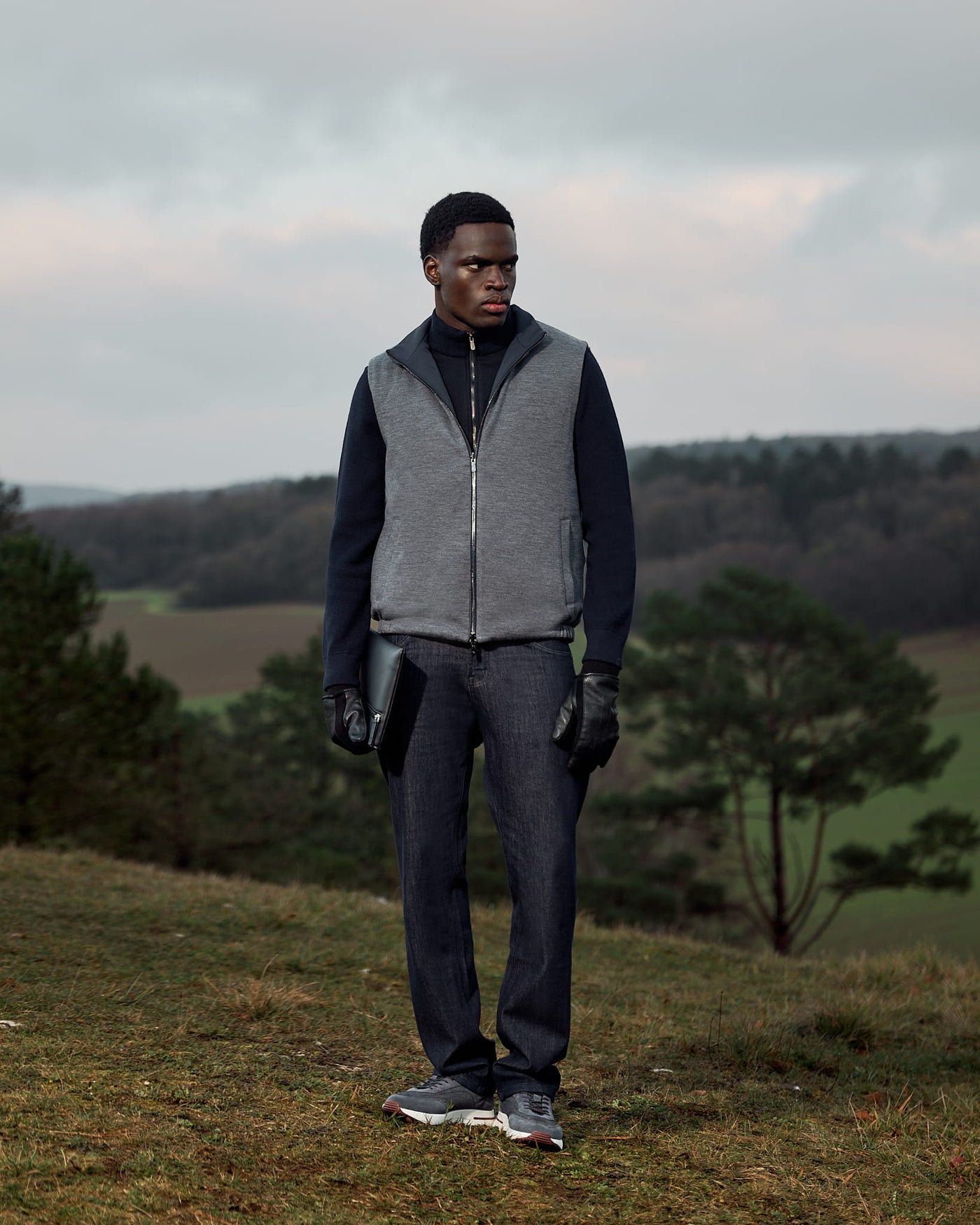 Person wearing a gray vest and dark pants standing in a field with trees and a cloudy sky.
