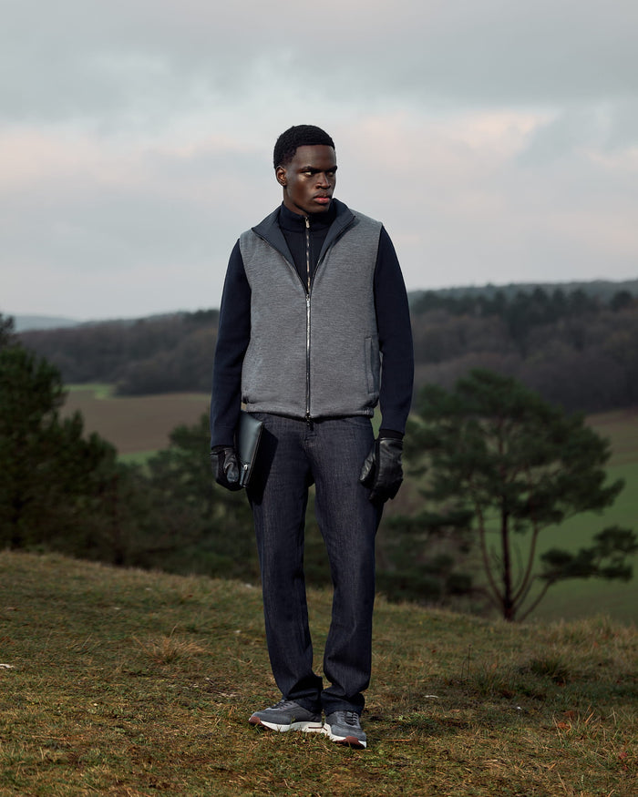 Person wearing a gray vest and dark pants standing in a field with trees and a cloudy sky.