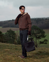 Man in brown jacket and dark pants holding a black bag in a field