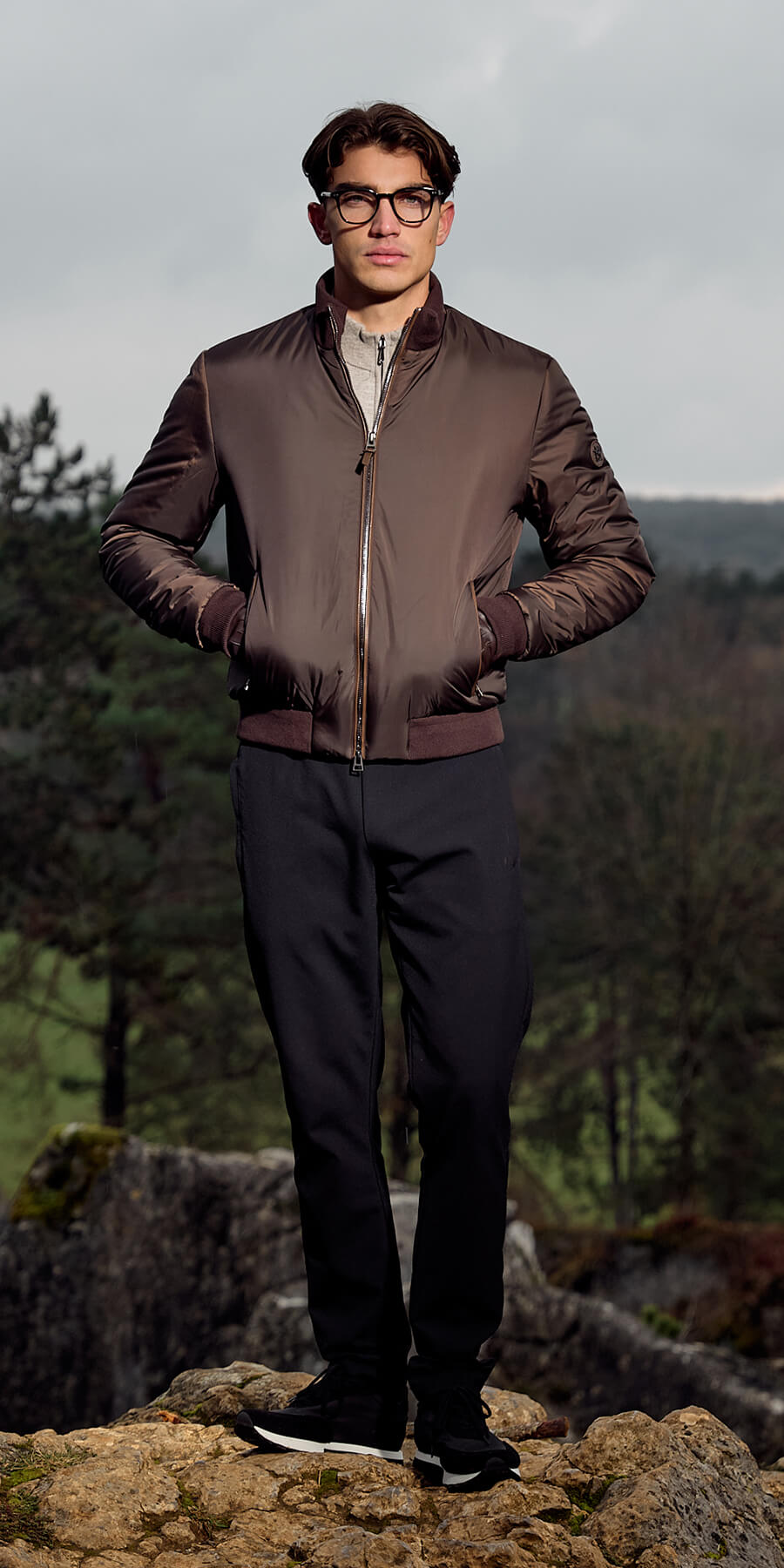 Man wearing a brown jacket and black pants standing on rocks with a forest background