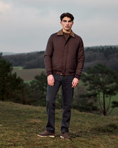 Man wearing a brown jacket and dark pants standing in a natural landscape.