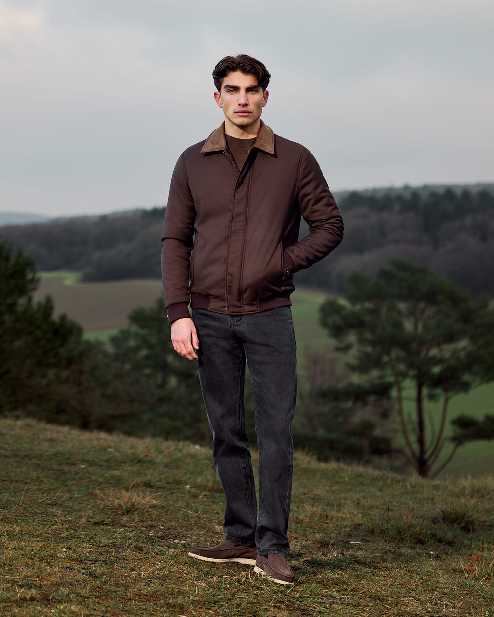 Man wearing a brown jacket standing in a field with trees and a cloudy sky in the background
