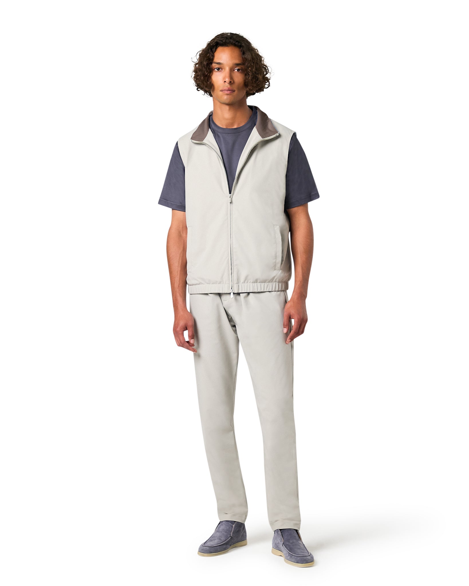 Man wearing a gray vest over a dark shirt with light pants on a white background