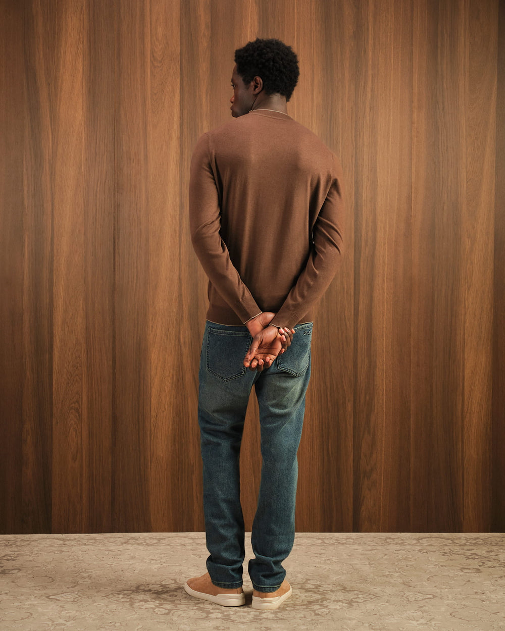Man wearing a brown sweater and blue jeans standing against a wooden paneled wall.