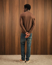Man wearing a brown sweater and blue jeans standing against a wooden paneled wall.