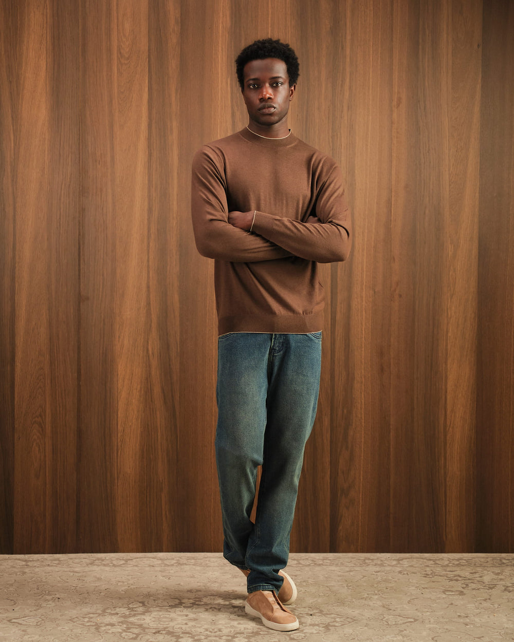 Man wearing a brown sweater and blue jeans standing against a wooden paneled wall.