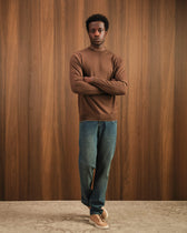Man wearing a brown sweater and blue jeans standing against a wooden paneled wall.