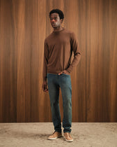 Man wearing a brown sweater and blue jeans standing against a wooden paneled wall.