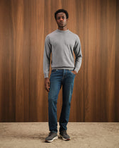 Man wearing a gray sweater and blue jeans standing against a wooden wall.