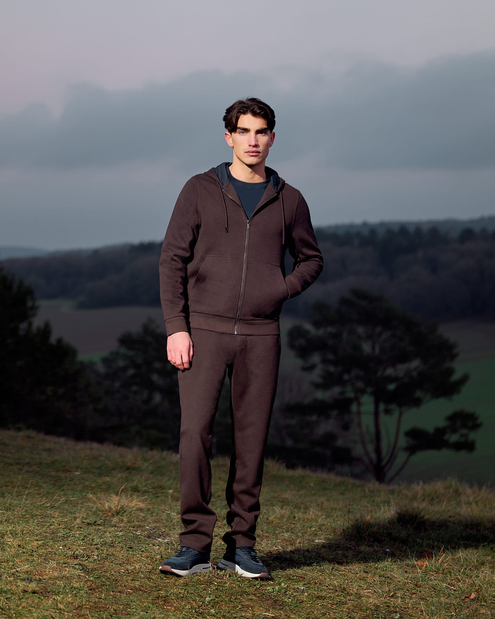 Man wearing a brown tracksuit standing in a field with trees and a cloudy sky in the background