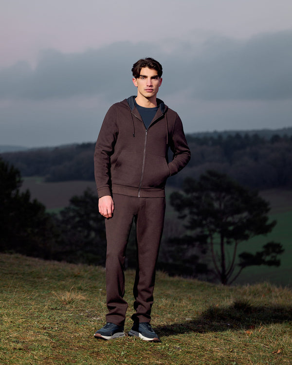Man wearing a brown tracksuit standing in a field with trees and a cloudy sky in the background