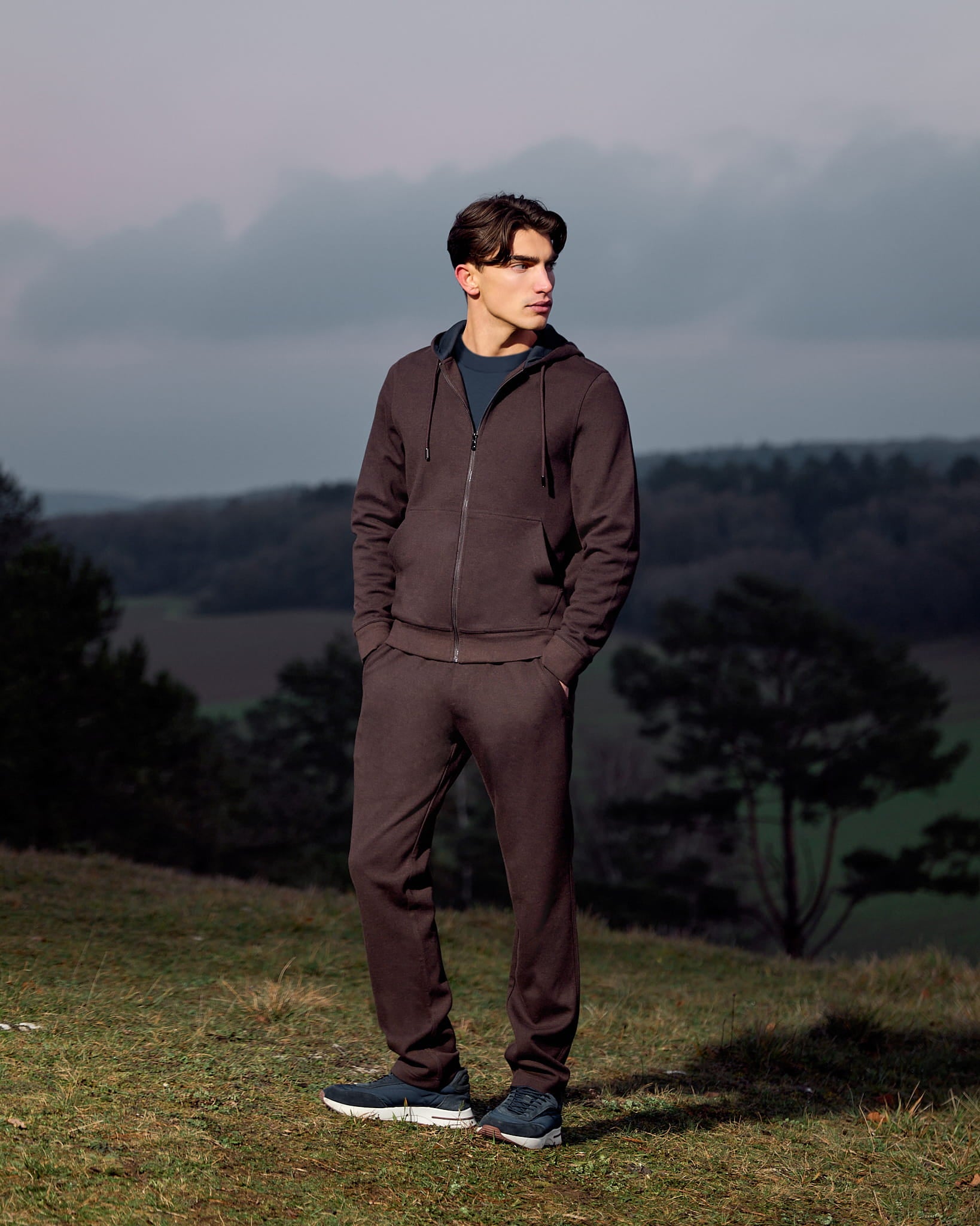 Man wearing a brown tracksuit standing in a field with trees and a cloudy sky in the background