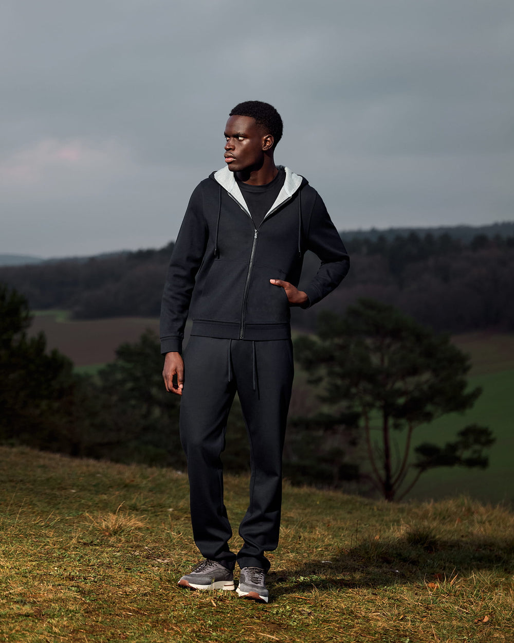 Man wearing a dark hoodie and pants standing in a field with trees and a cloudy sky in the background