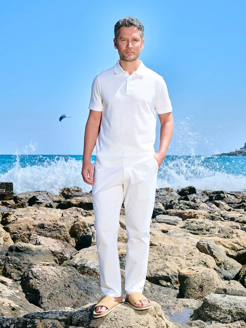 Man in an ivory polo and white trousers on a rocky shore
