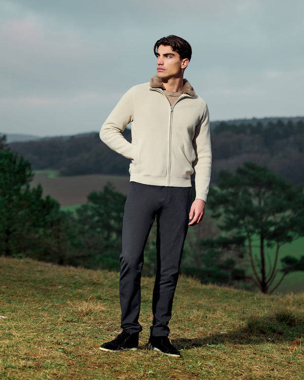 Man wearing a beige zip-up hoodie and dark pants standing in a scenic outdoor setting with trees and mountains.