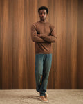 Man wearing a brown sweater and blue jeans standing against a wooden paneled wall.