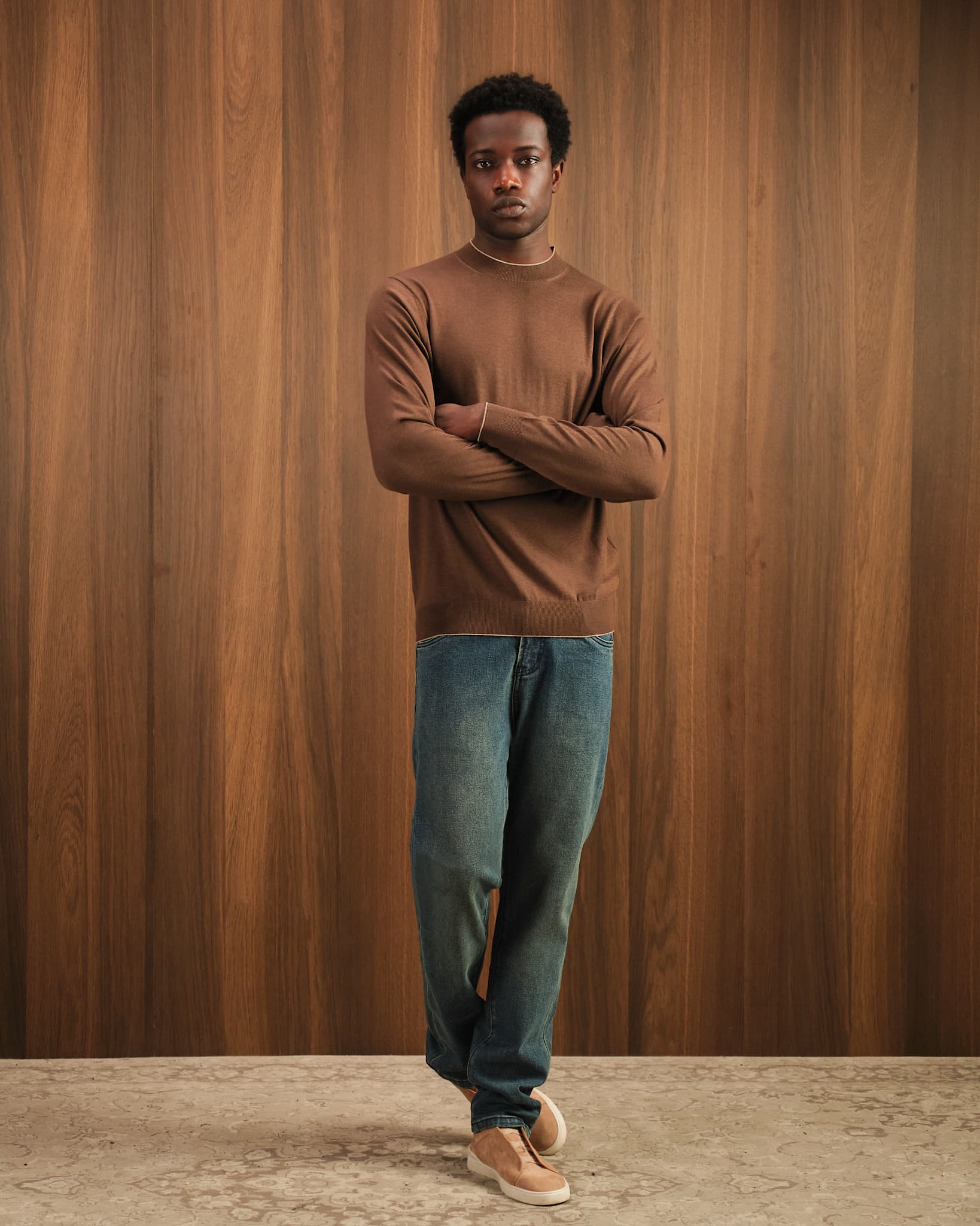 Man wearing a brown sweater and blue jeans standing against a wooden paneled wall.