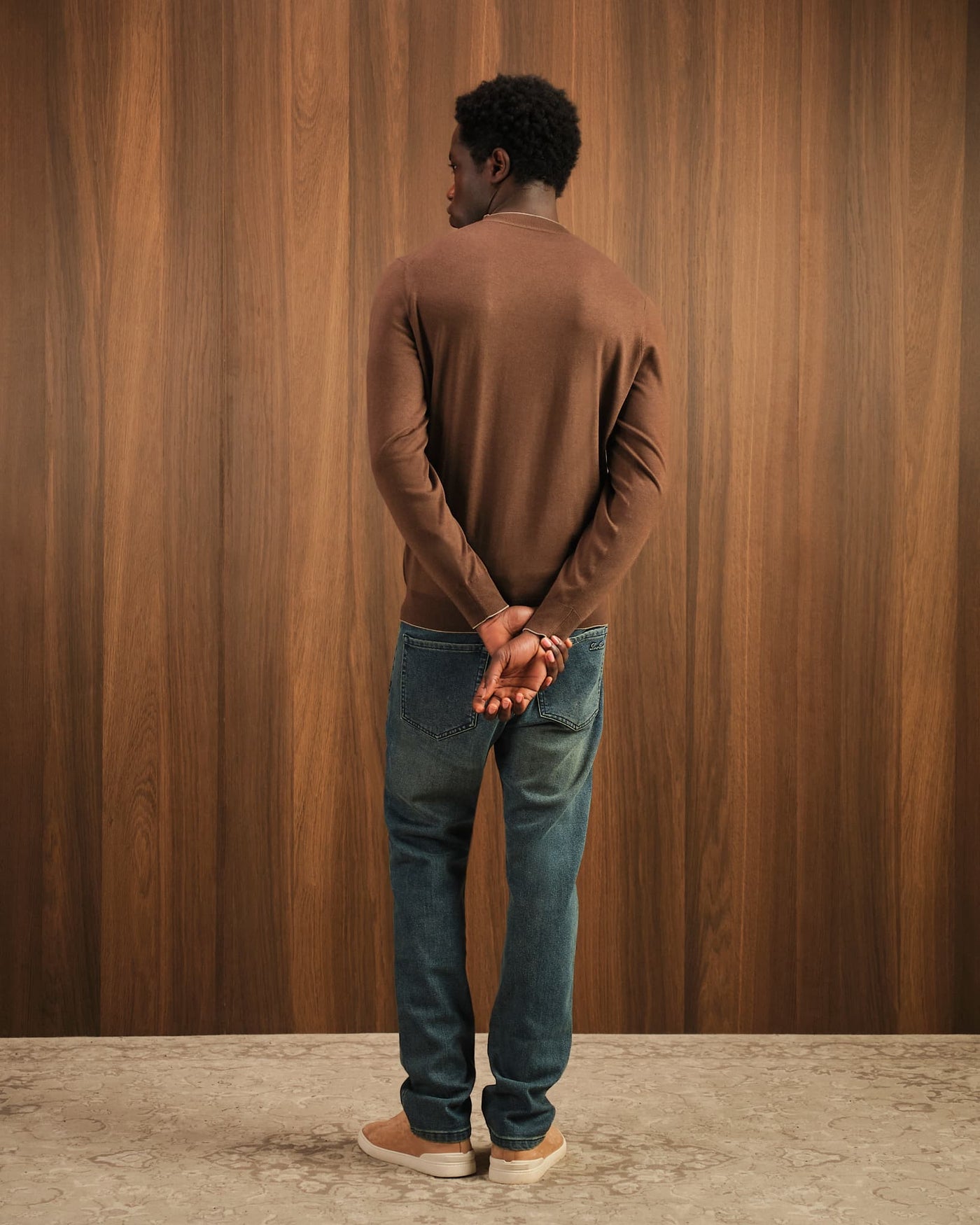 Man wearing a brown sweater and blue jeans standing against a wooden paneled wall.
