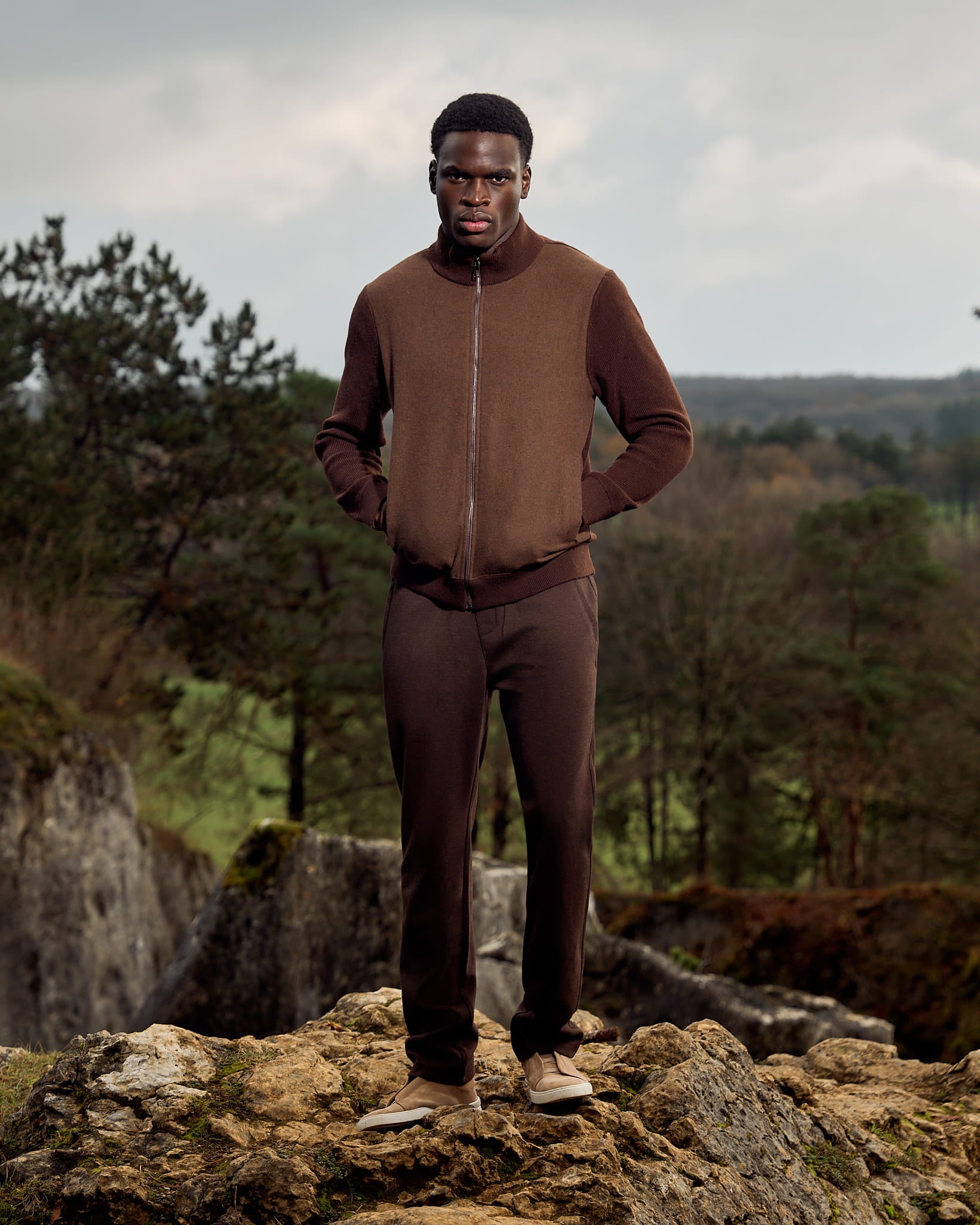 Man in brown outfit standing on a rocky outcrop with a natural landscape in the background
