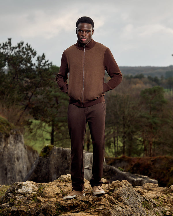 Man in brown outfit standing on a rocky outcrop with a natural landscape in the background