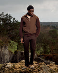 Man in brown outfit standing on a rocky outcrop with a forest background