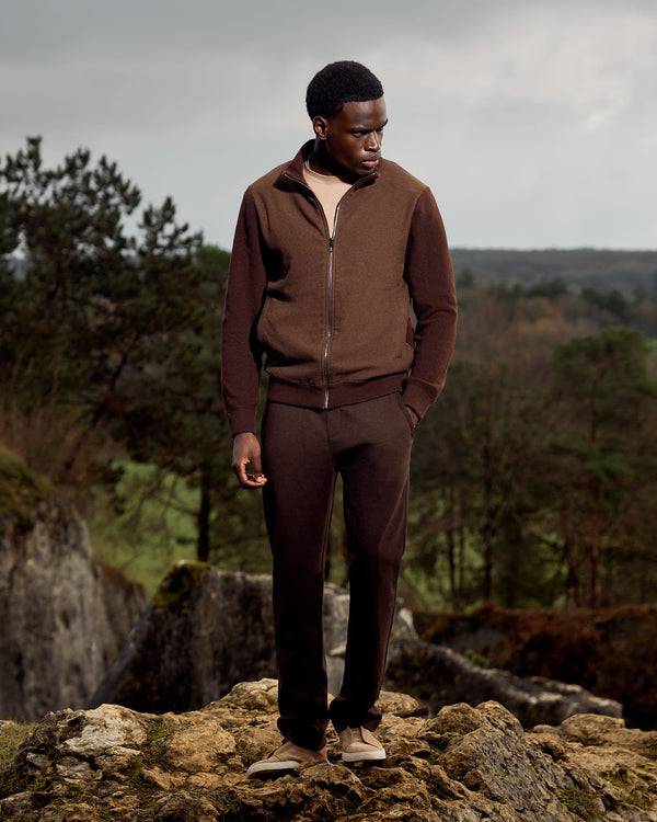 Man in brown outfit standing on a rocky outcrop with a forest background