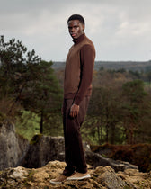 Man in brown outfit standing on rocky terrain with a forest background