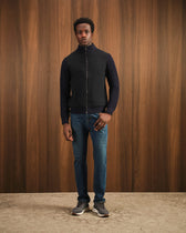 Man wearing a black zip-up jacket and blue jeans standing against a wooden paneled wall.