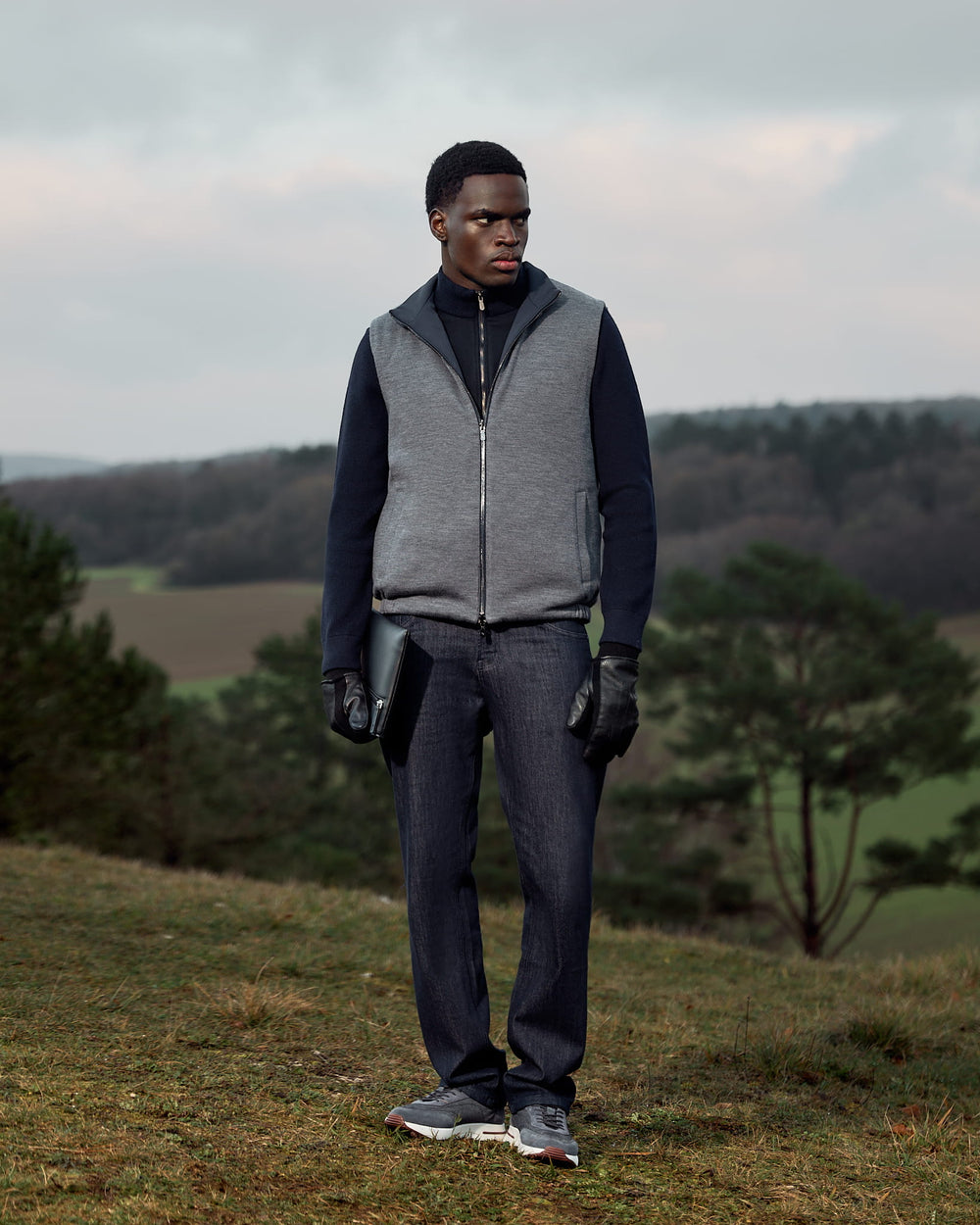 Man wearing a gray vest and dark pants standing in a field with trees and sky in the background
