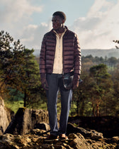 Man wearing a brown puffer jacket standing on rocky terrain with trees and mountains in the background