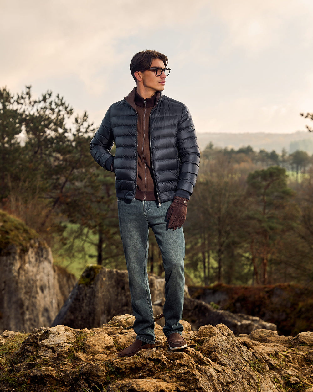Man wearing a blue puffer jacket standing on a rocky outcrop with trees and mountains in the background.