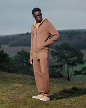 Man wearing a brown tracksuit standing in a natural setting with trees and grass.