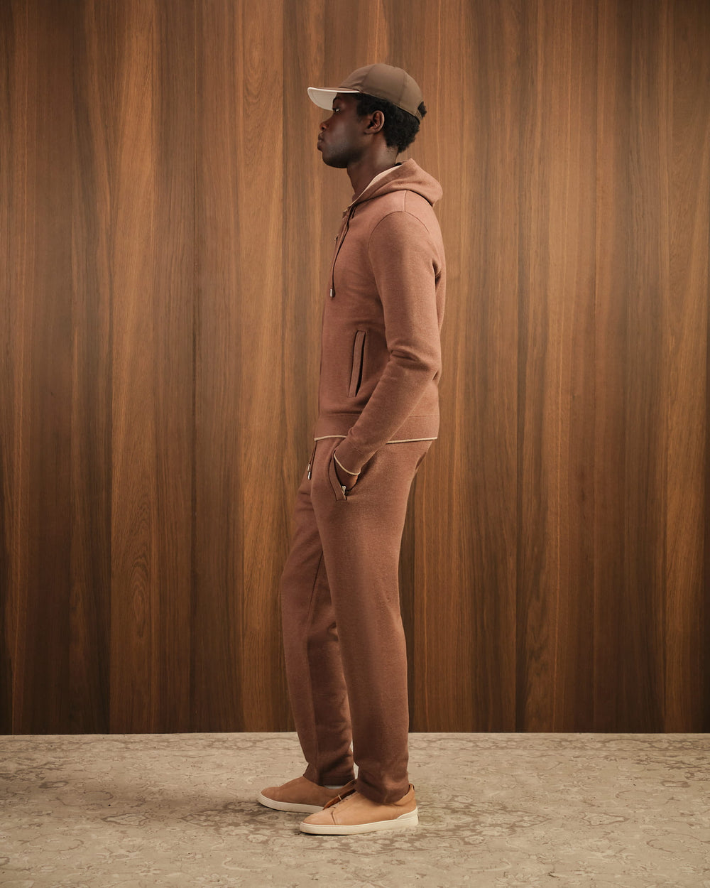 Man wearing a brown tracksuit and cap against a wooden paneled wall.