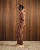 Man wearing a brown tracksuit and cap against a wooden paneled wall.