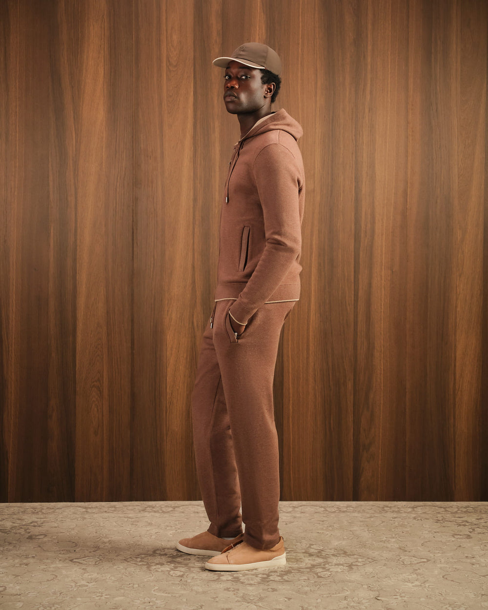 Man wearing a brown tracksuit and cap against a wooden paneled wall.