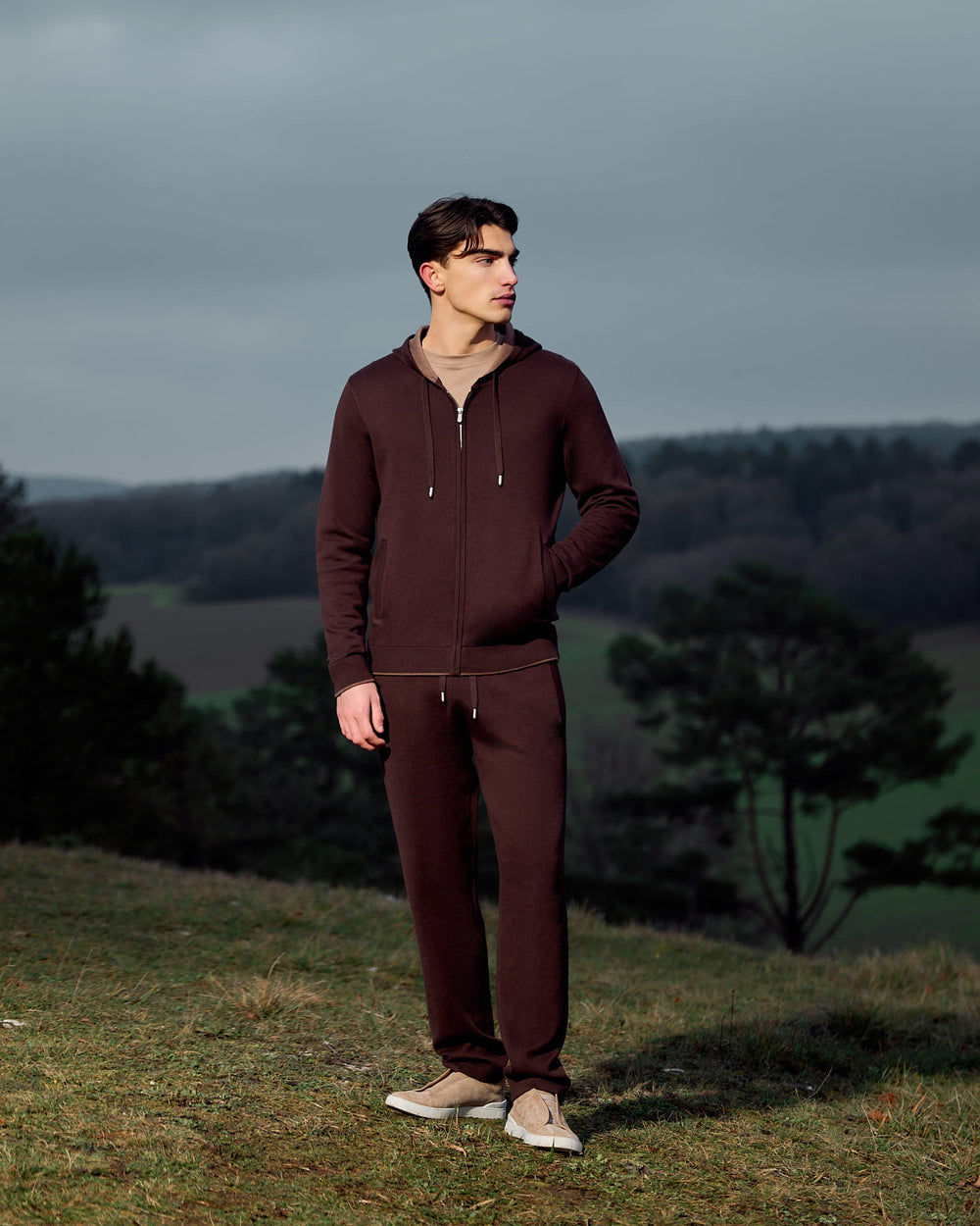 Man wearing a brown tracksuit standing on a grassy hill with a scenic background