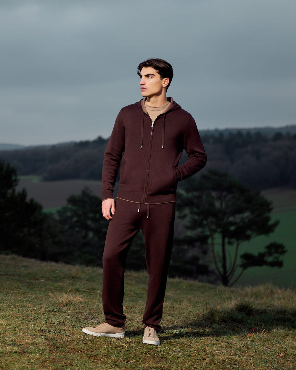 Man wearing a maroon tracksuit standing in a field with a scenic background
