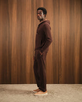 Man wearing a brown hoodie and pants standing against a wooden paneled wall.