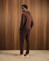 Man wearing a brown tracksuit standing against a wooden paneled wall.