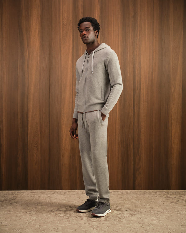 Man wearing a gray hoodie and pants standing against a wooden paneled wall.