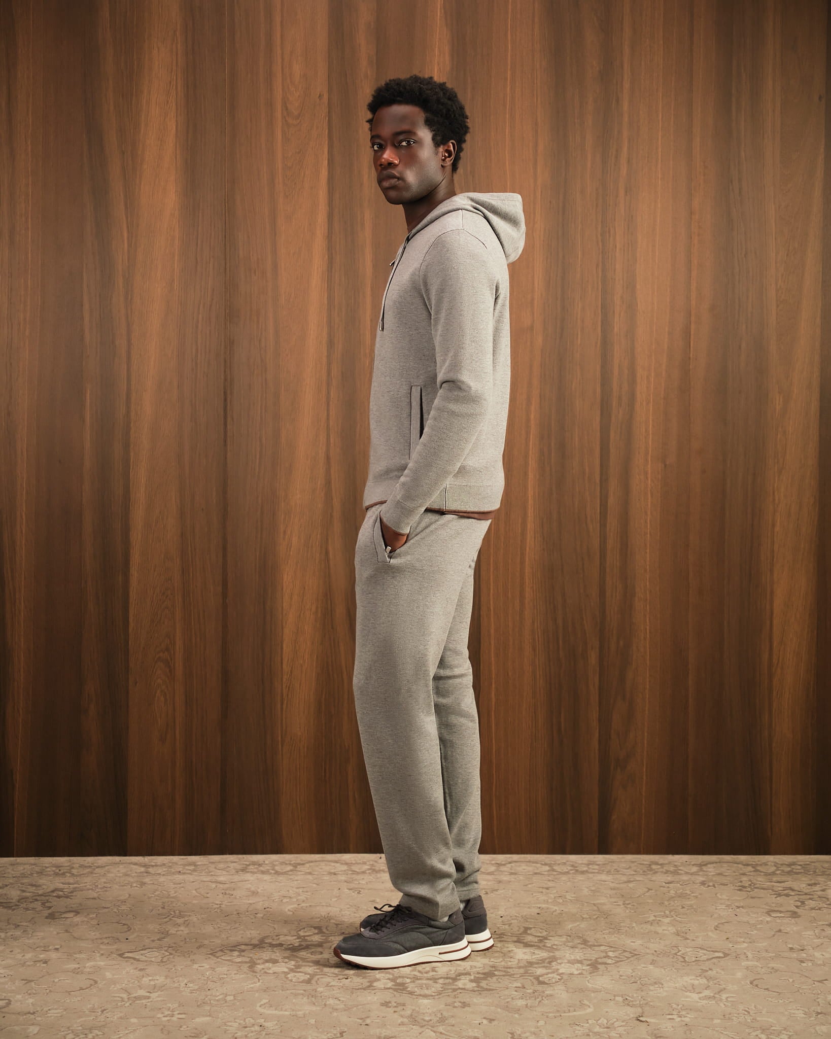 Man wearing a gray hoodie and pants standing against a wooden paneled wall.