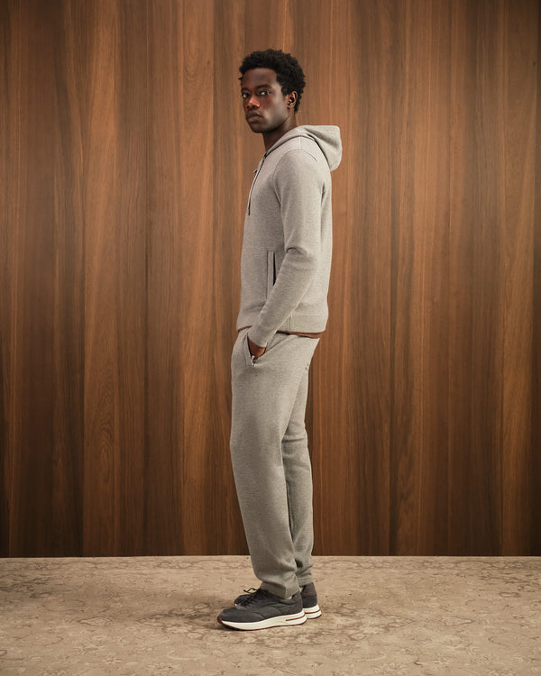 Man wearing a gray hoodie and pants standing against a wooden paneled wall.