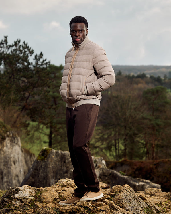 Man wearing a beige puffer jacket and brown pants standing on rocky terrain with trees and mountains in the background.