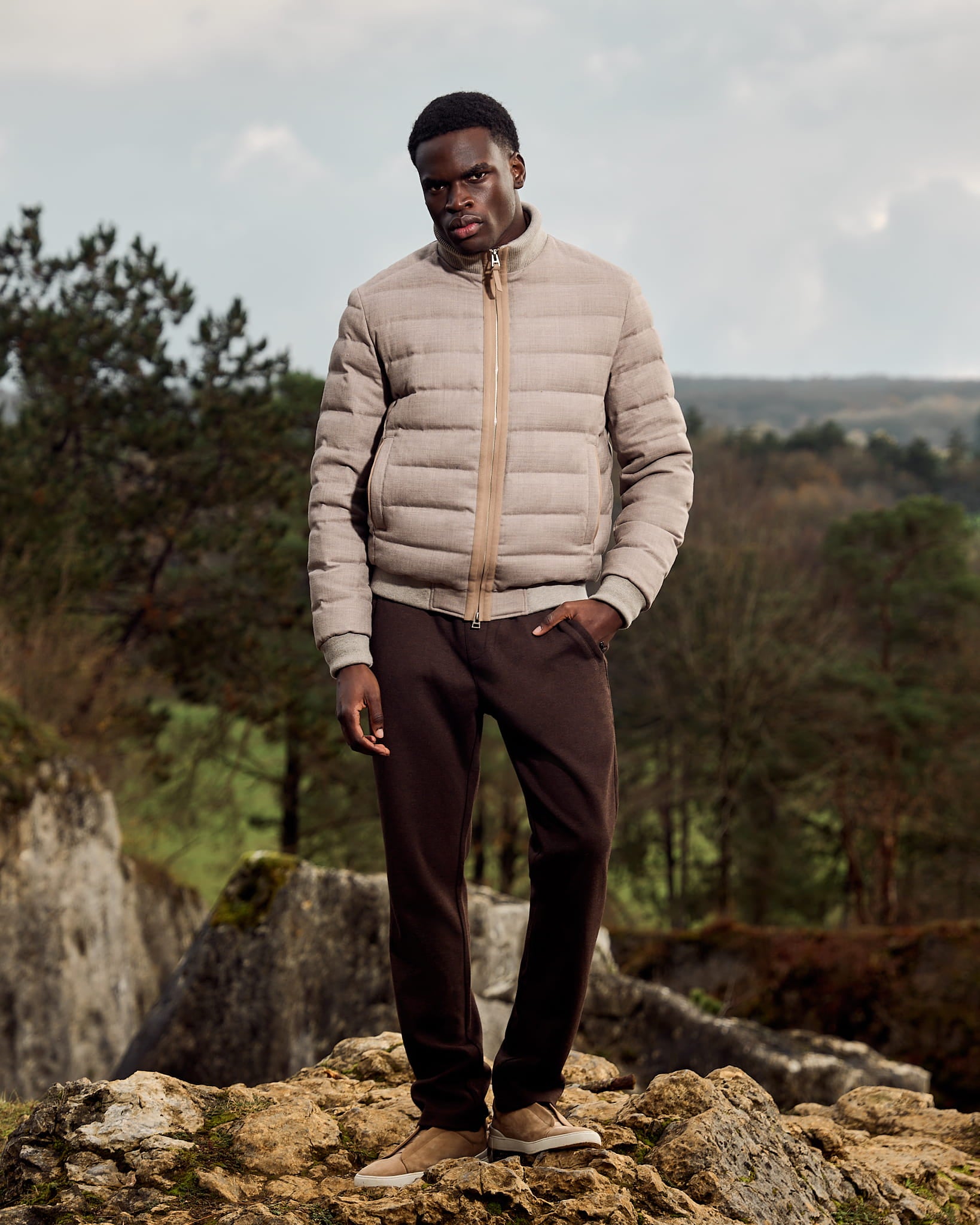 Man wearing a beige puffer jacket and brown pants standing on rocks with a natural landscape in the background