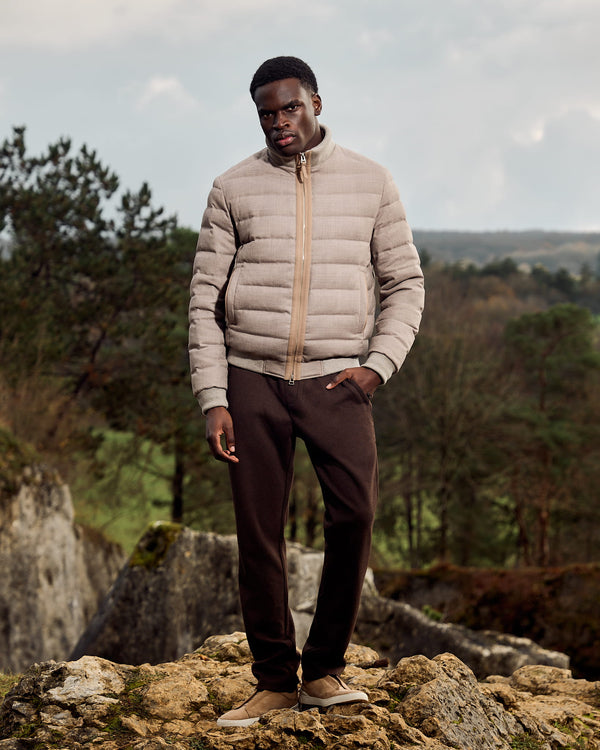 Man wearing a beige puffer jacket and brown pants standing on rocks with a natural landscape in the background