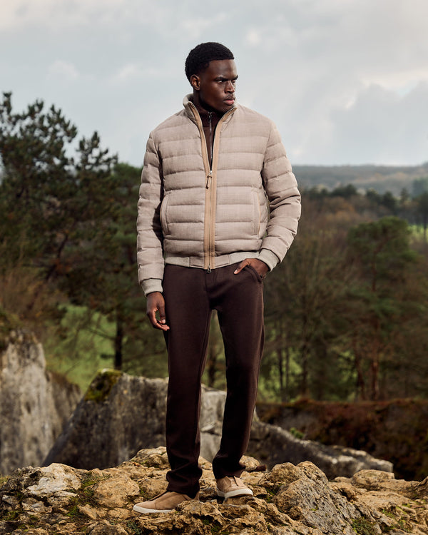 Man wearing a beige puffer jacket and dark pants standing on rocky terrain with trees and mountains in the background.