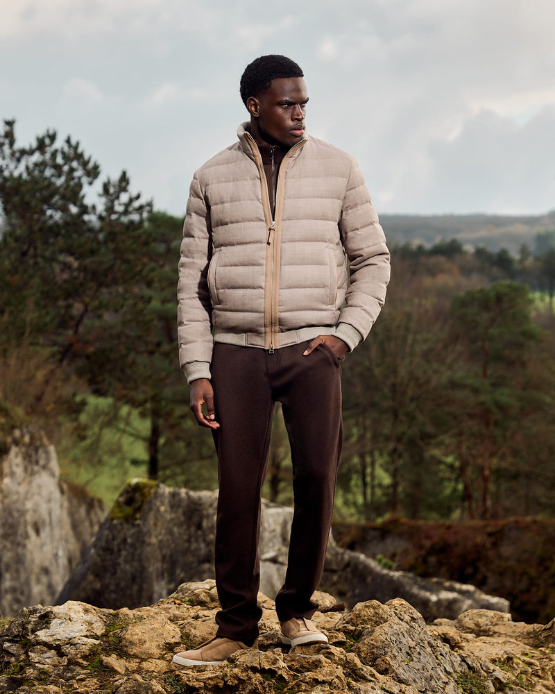 Man wearing a beige puffer jacket and dark pants standing on rocky terrain with trees and mountains in the background.