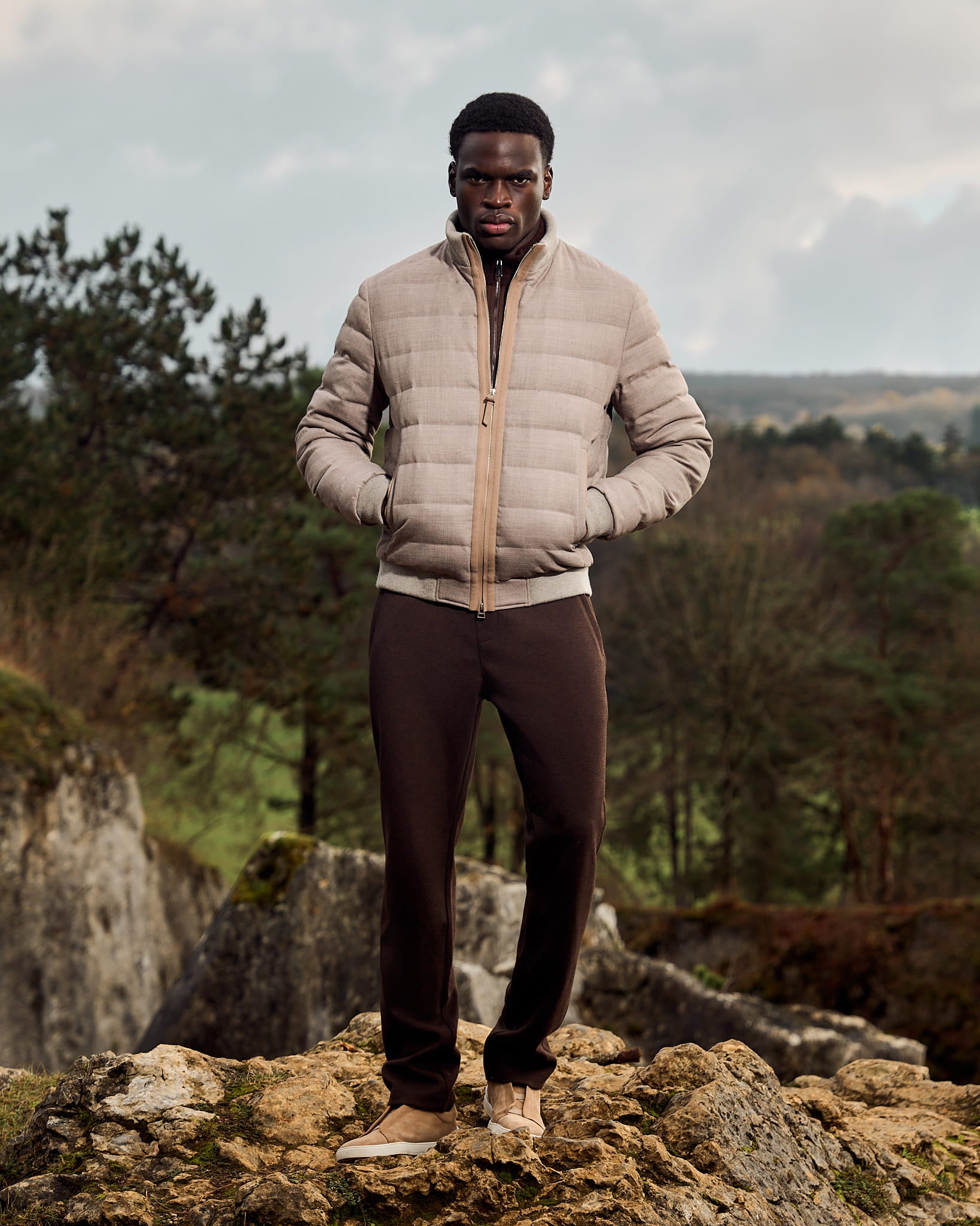 Man wearing a beige puffer jacket and dark pants standing on rocky terrain with trees and sky in the background